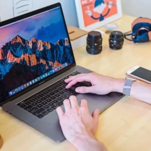 A person typing on a laptop with a mountain wallpaper. A smartphone, headphones, camera lenses, and a mug are on the desk. A person typing on a laptop at a desk. The screen shows a mountain wallpaper. Nearby, there are headphones, camera lenses, a smartphone, and a coffee mug.