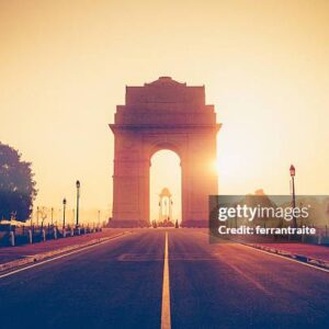 A large stone archway stands at the end of an empty road, with the sun setting behind it, casting a warm glow. India Gate at sunrise with an empty road leading to the monument.