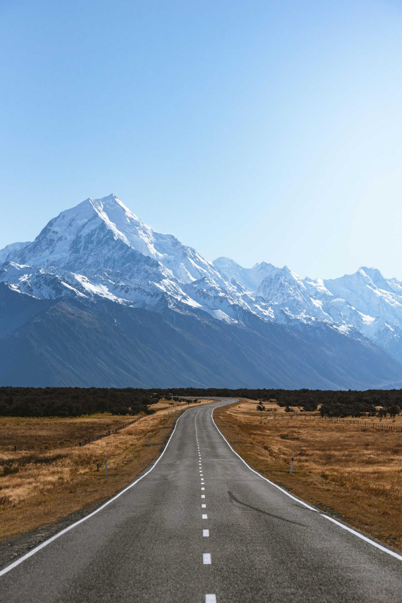 A straight empty road leading towards a range of snow-capped mountains under a clear blue sky. A long, straight road leading towards snow-capped mountains under a clear blue sky.
